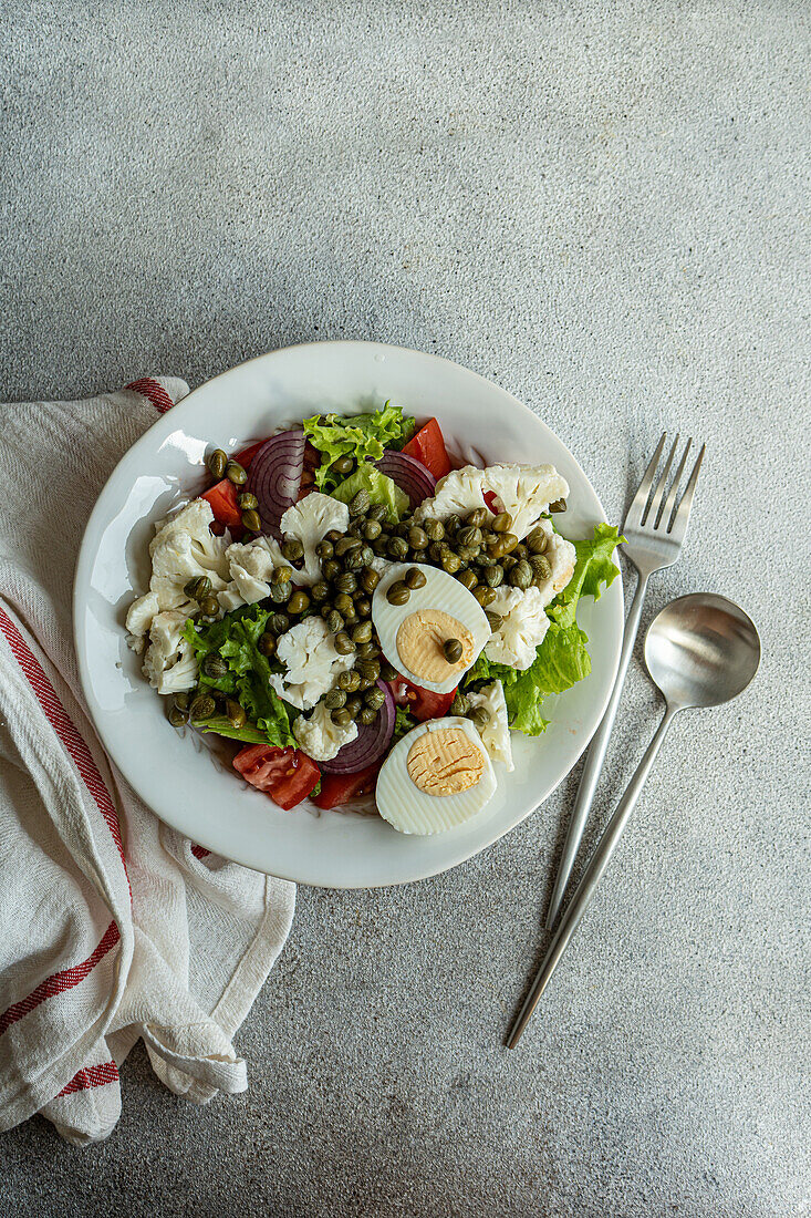 Top view of plate of Keto salad with salad leaves, boiled eggs, capers, tomato, red onion and cauliflower vegetables placed on table between napkin and cutlery