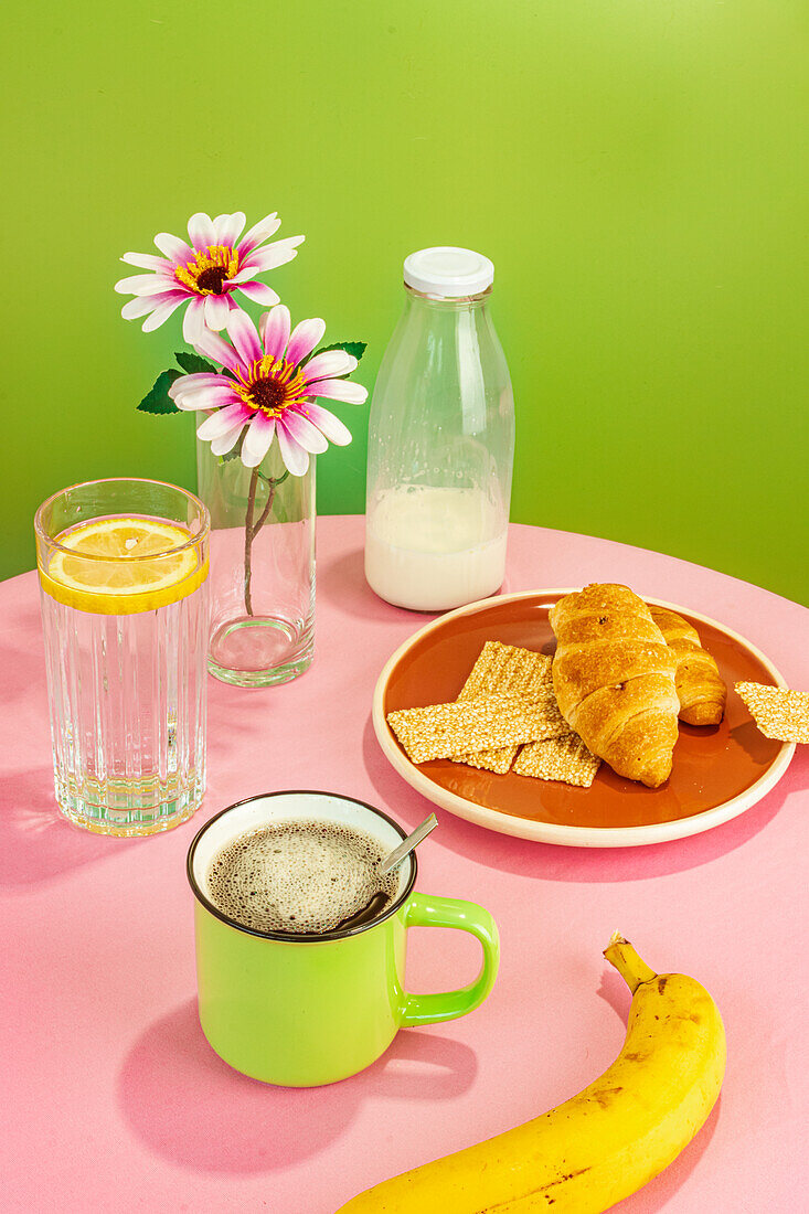 Appetitanregende Croissants und Banane neben Glasflasche mit Milch und Wasser mit Zitronenscheibe vor Blumen in Vase und Tasse mit aromatischem Kaffee auf rosa Tisch vor grünem Hintergrund