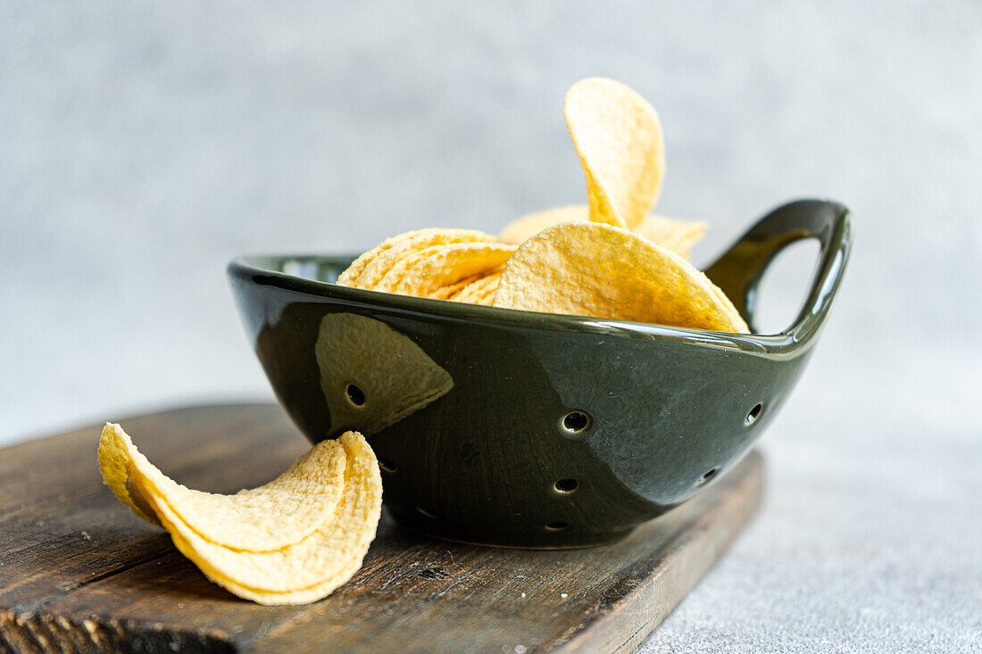 Stacked pieces of delicious crispy yellow potato chips in black bowl over wooden table in bright room against blurred background