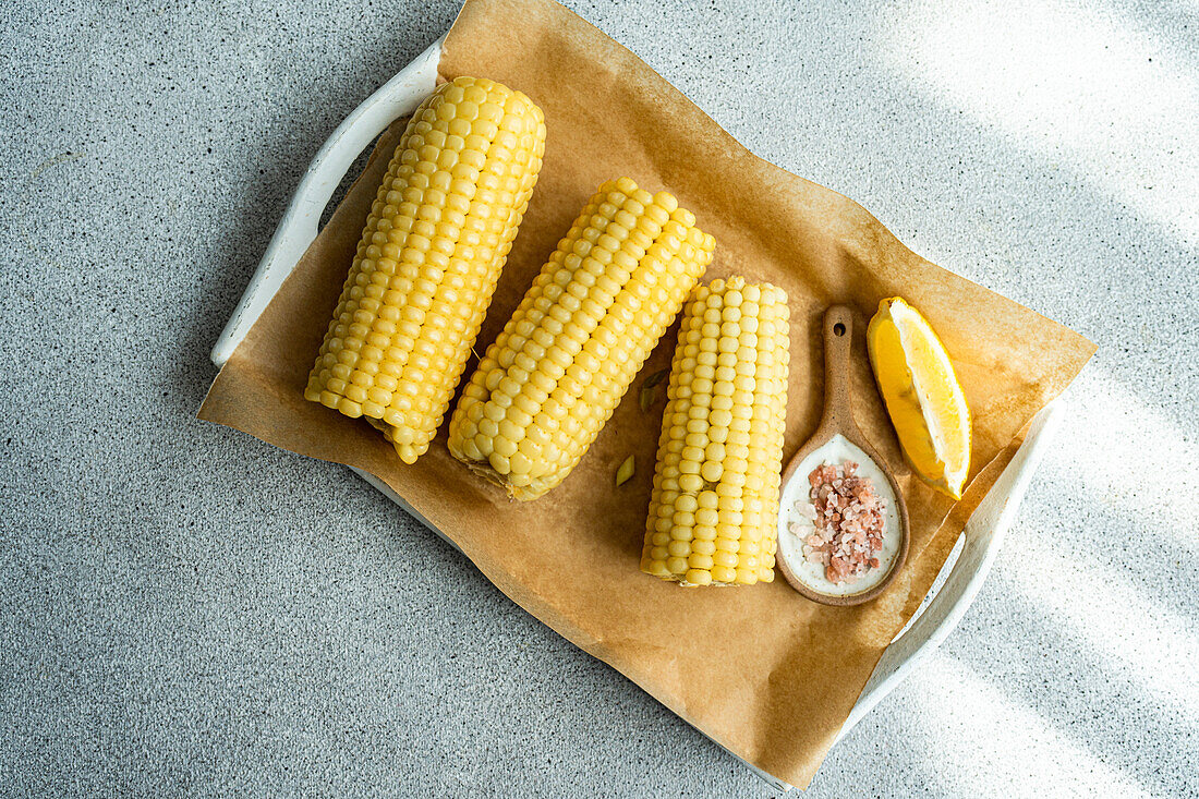 Top view of wooden chopping board with ripe cooked corncobs placed on concrete table in kitchen with bowl with sea salt and napkin