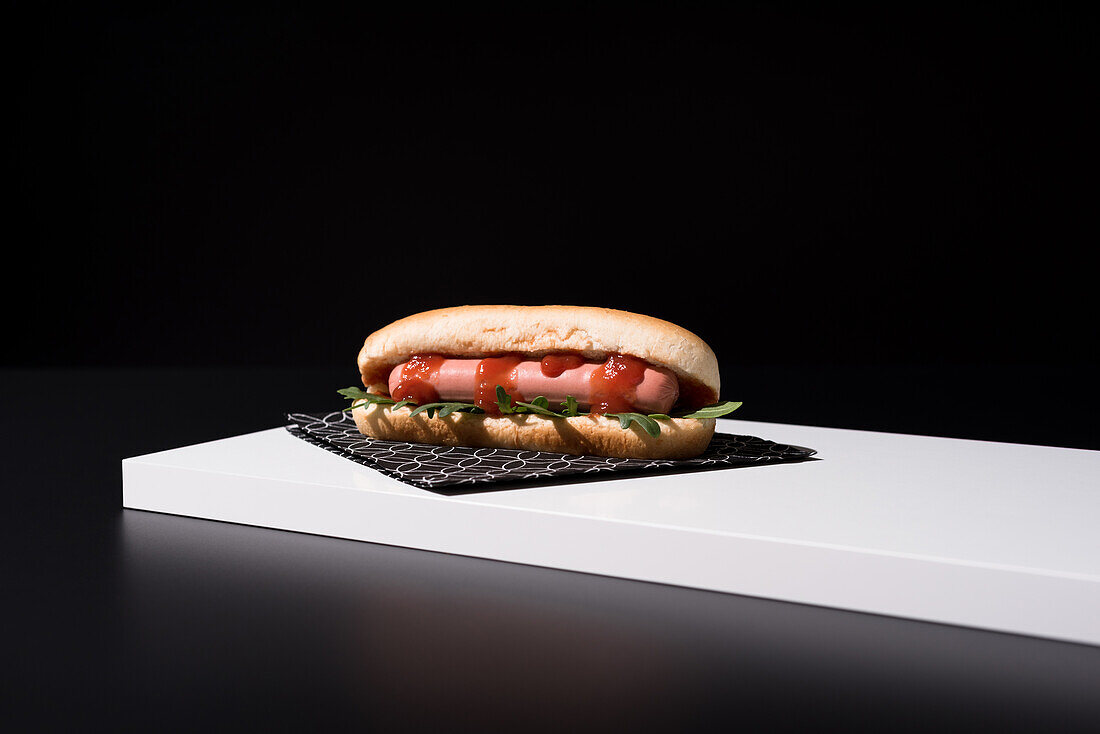 Appetizing bun with sausage and ketchup with rocket salad served table mat over white wooden board against black background in studio