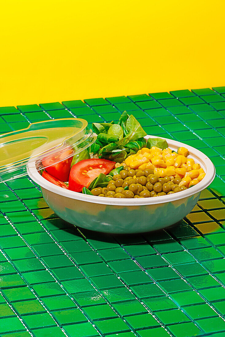 Salad bowl with slices of tomato, spinach leaves, corn kernels and peas placed on green surface against yellow wall