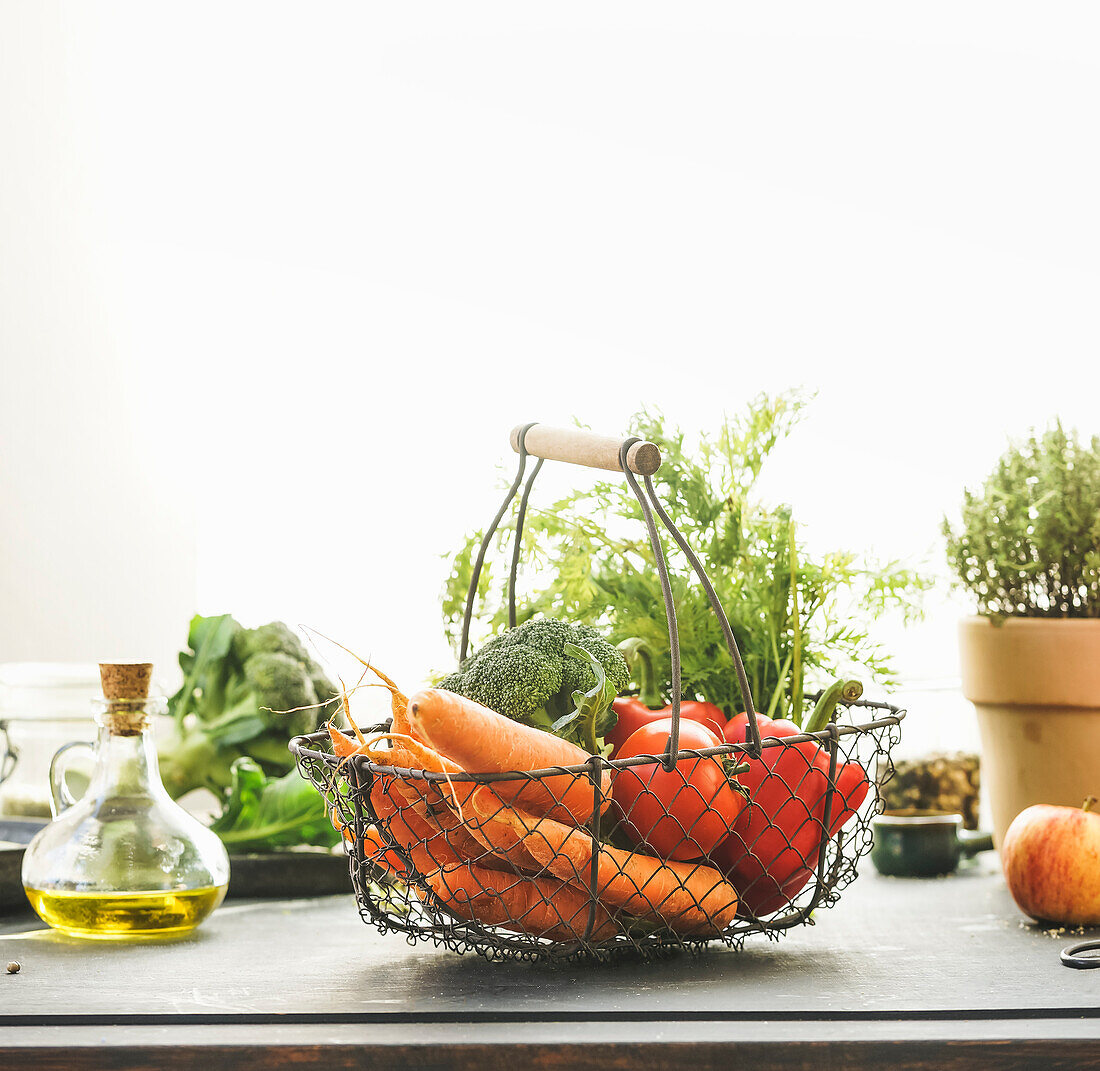 Lebensmittelkorb aus Metall mit Bio-Gemüse: Karotten, Brokkoli, Paprika und Tomaten auf einem Tisch mit anderen gesunden Zutaten vor einem Fenster mit Sonnenlicht. Gesunde Lebensweise. Vorderansicht