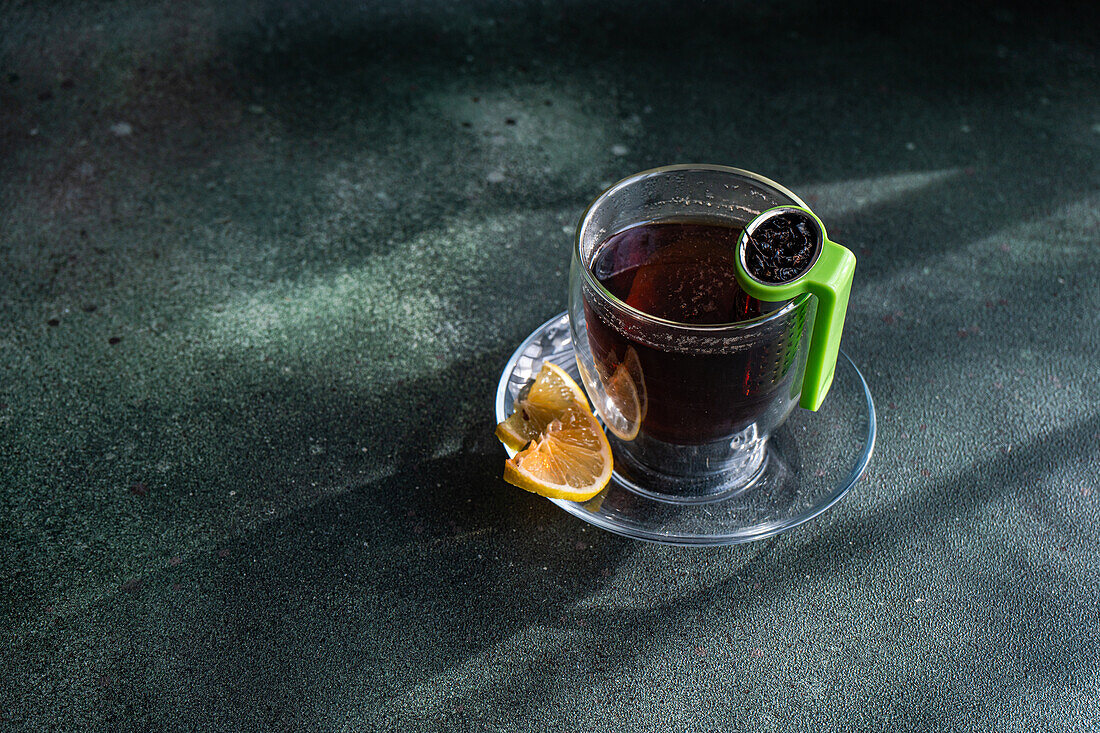 From above of clear glass of tea with a slice of lemon and a green tea strainer on a textured dark background