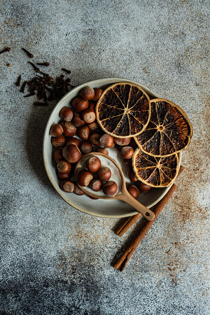From above of fresh nuts placed in ceramic bowl with dried orange peels, cinnamon and cloves a concrete background