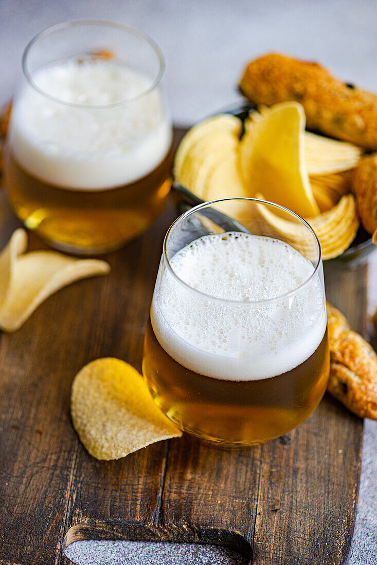 High angle of tasty pieces of crisp potato chips in bowl with bread sticks and glasses of beer on wooden board placed over table in daylight