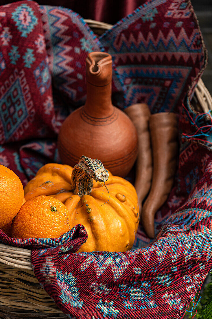 A rustic display featuring a clay pot, vibrant oranges, a pumpkin and traditional woven fabrics with intricate patterns in shades of red and blue inside a basket.