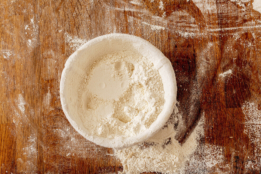 Overhead view of bowl with flour placed on messy wooden table in kitchen of bakehouse during cooking process