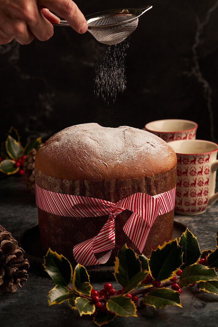 Close-up of a hand sprinkling powdered sugar over a festive panettone, surrounded by holiday decorations and ceramic mugs