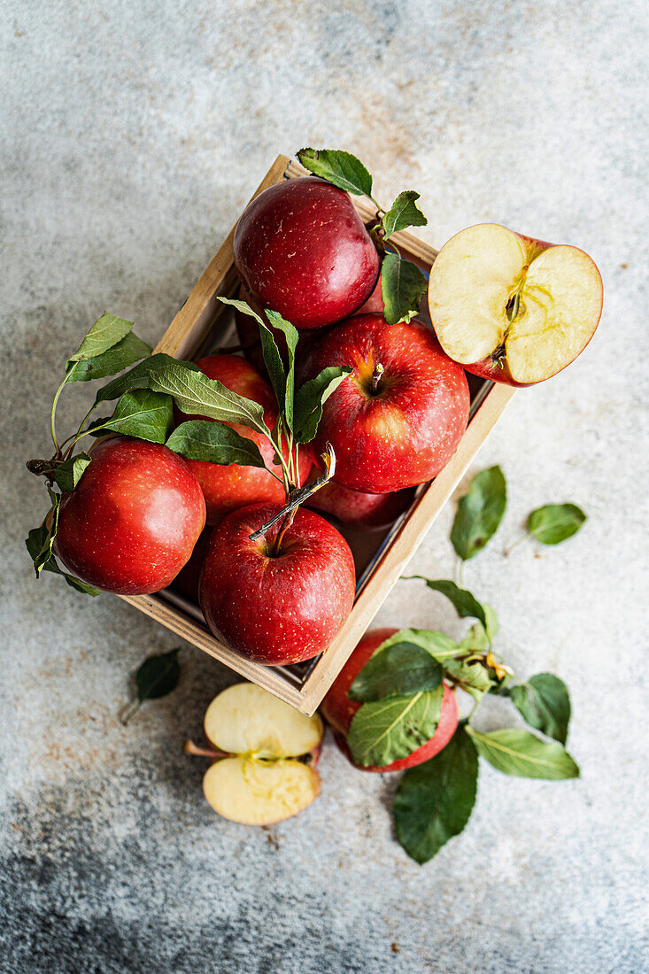 Draufsicht auf ganze frische, rote, reife, köstliche Bio-Äpfel mit grünen Blättern, die in eine Holzkiste mit halbierten Stücken gefüllt und auf eine graue Fläche im Tageslicht gelegt wurden