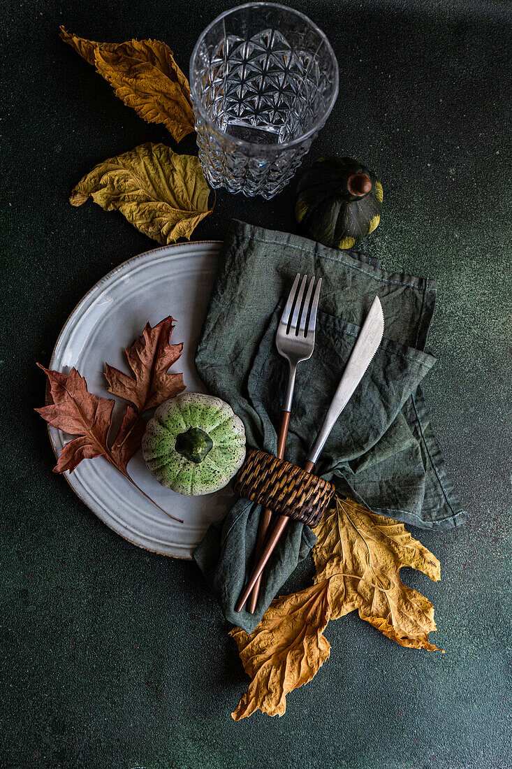 Draufsicht auf einen herbstlich gedeckten Tisch mit Serviette, Messer und Gabel auf einem Teller neben bunten Blättern, kleinen Kürbissen und einem leeren Glas auf dunkler Oberfläche