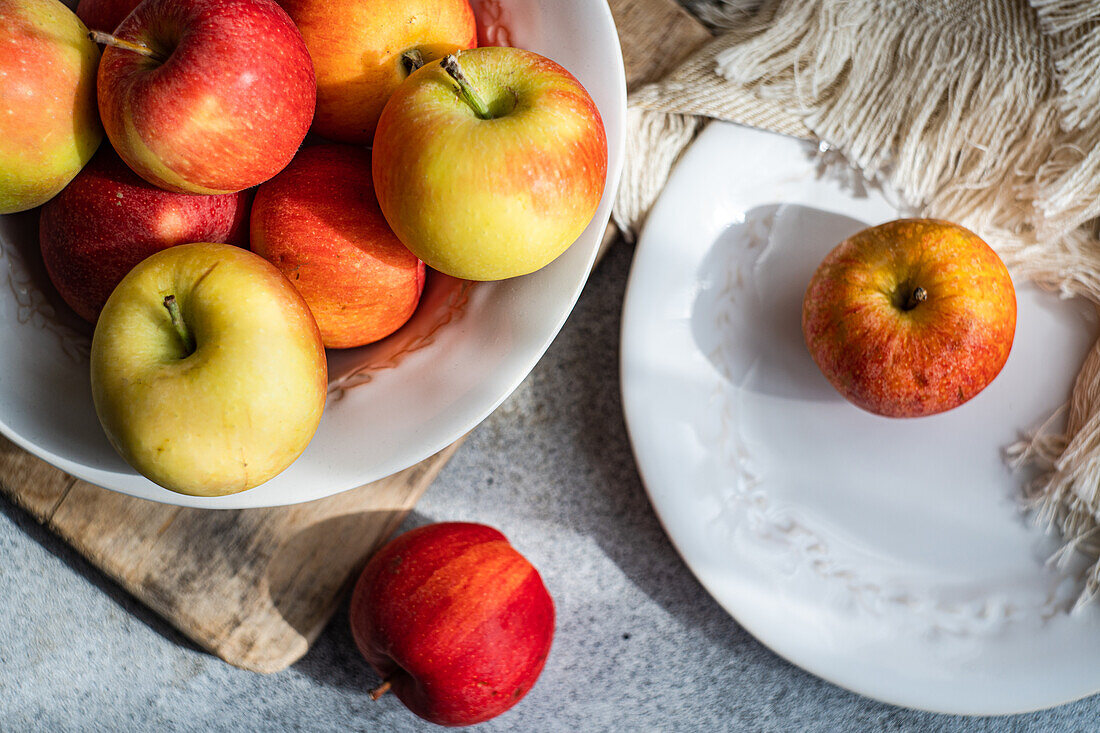 From below of collection of ripe, colorful apples presented on a rustic wooden board, with a white plate and draped fabric creating a serene kitchen setting