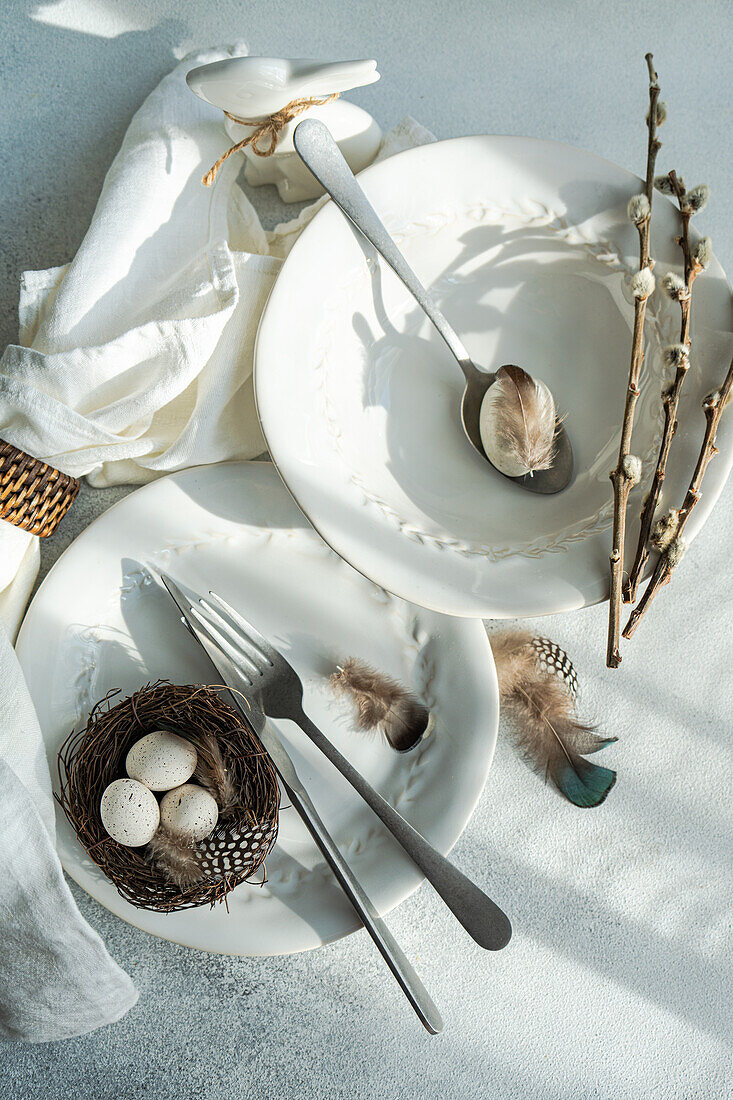 Draufsicht auf ein elegantes Osterspeisenarrangement mit makellos weißen Tellern mit zartem Prägemuster und einem gewebten Nest mit gesprenkelten Eiern auf einem Teller, begleitet von Gabel und Messer, Federn und Zweigen