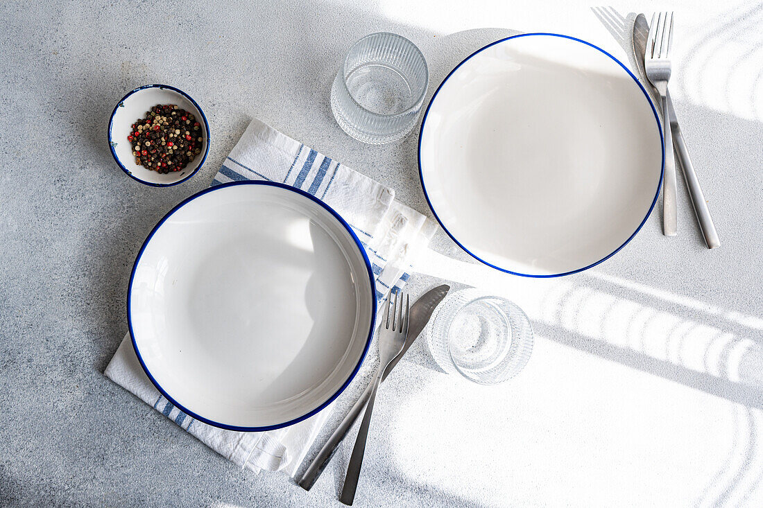Modernes Geschirr auf einer Marmorfläche, darunter weiße Teller mit blauem Rand, Silberbesteck und eine gestreifte Serviette, die eine elegante Tischdekoration bilden