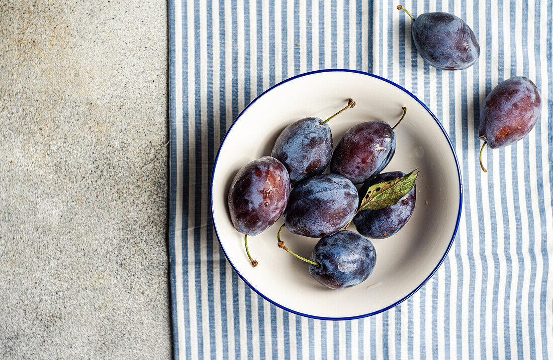 Top view of ripe plums served in ceramic bowl on striped napkin against gray surface