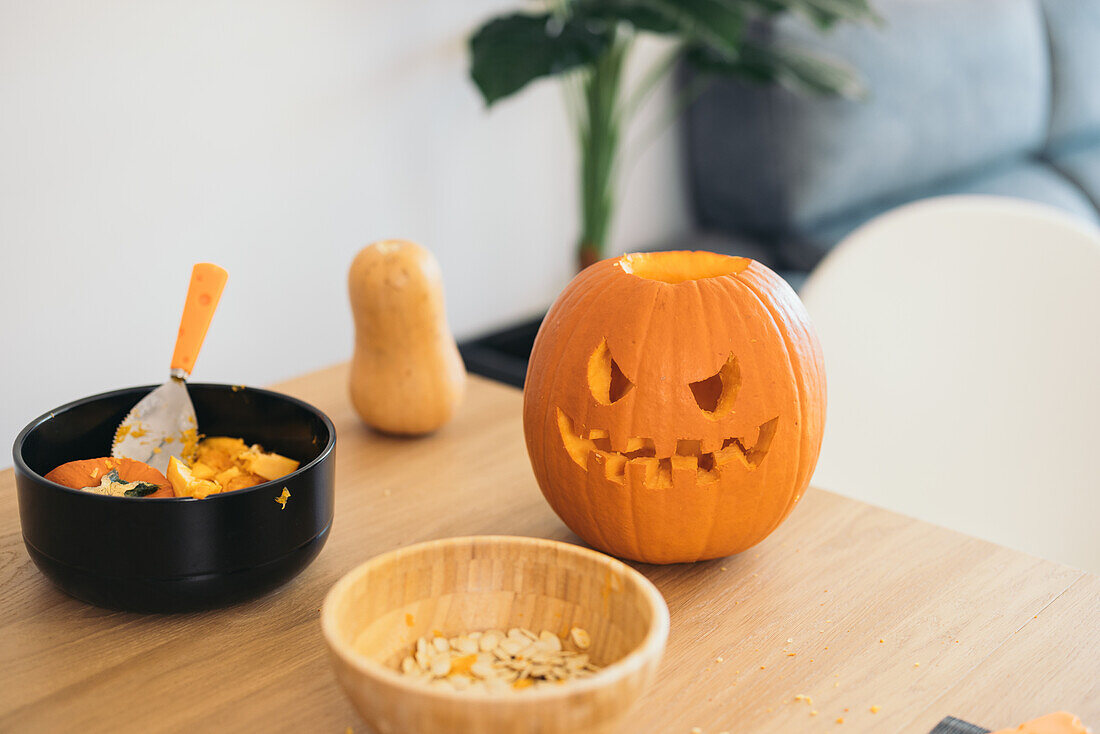 Orange pumpkin carved and ready for Halloween occasion and bowls with seeds and pulp placed on wooden table against blurred sofa set and potted plants