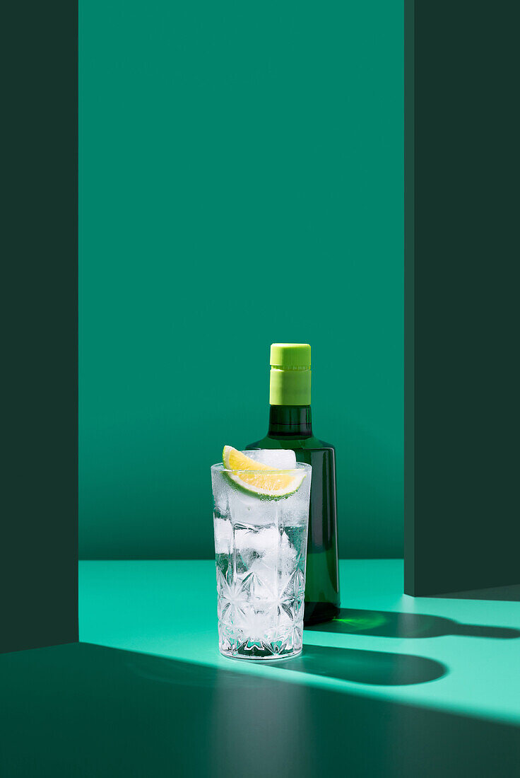 A minimalist composition featuring a gin bottle behind a glass of tonic water adorned with a lime slice, all against a two-tone green background.