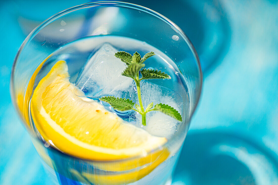 High angle of Summer cocktail with lemon vodka, slice of lemon and wild mint leaves served with ice placed on blue table