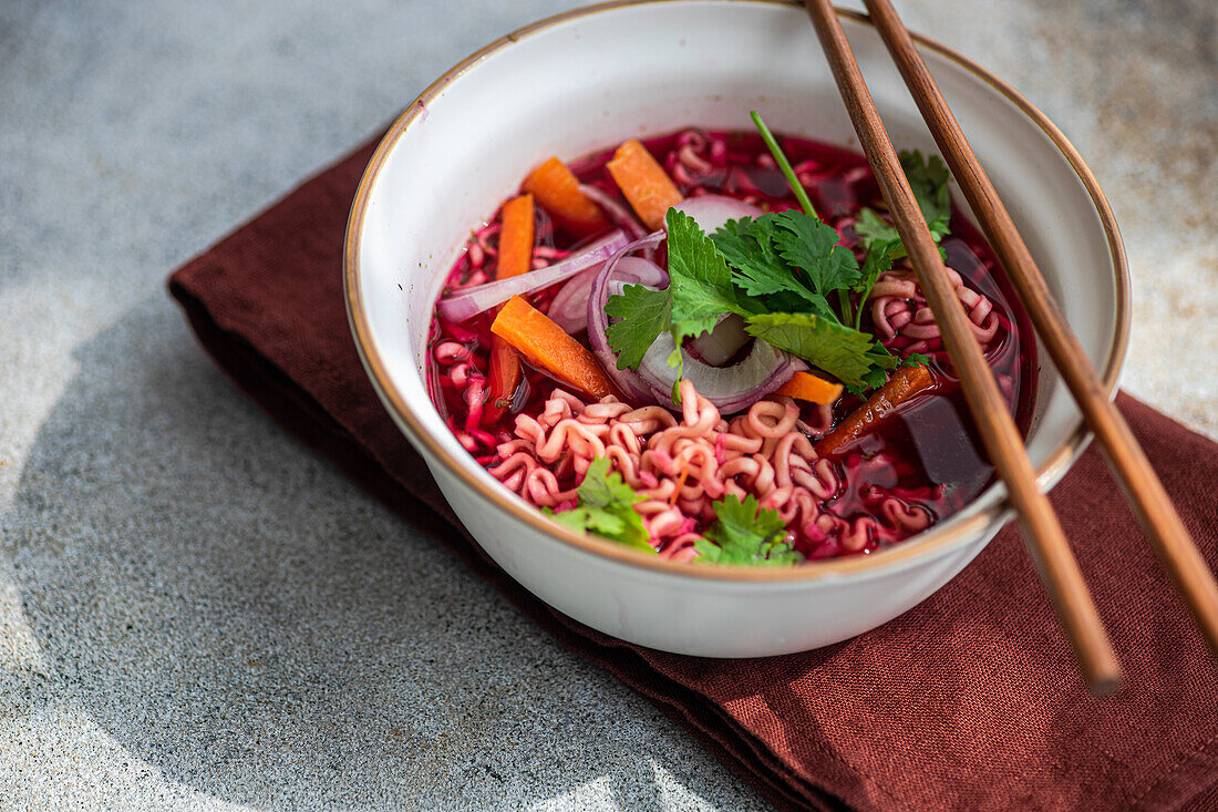 Closeup of bowl with beetroot soup with onion, coriander and noodles in Asian style served on bowl and chopsticks on napkin against gray surface in daylight