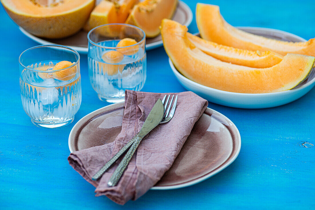 In Scheiben geschnittene frische Bio-Orangen-Moschus-Melone und Melonenkugeln in transparenten Gläsern mit Eiswürfeln, serviert auf einem Tisch mit Serviette, Gabel und Messer bei Tageslicht