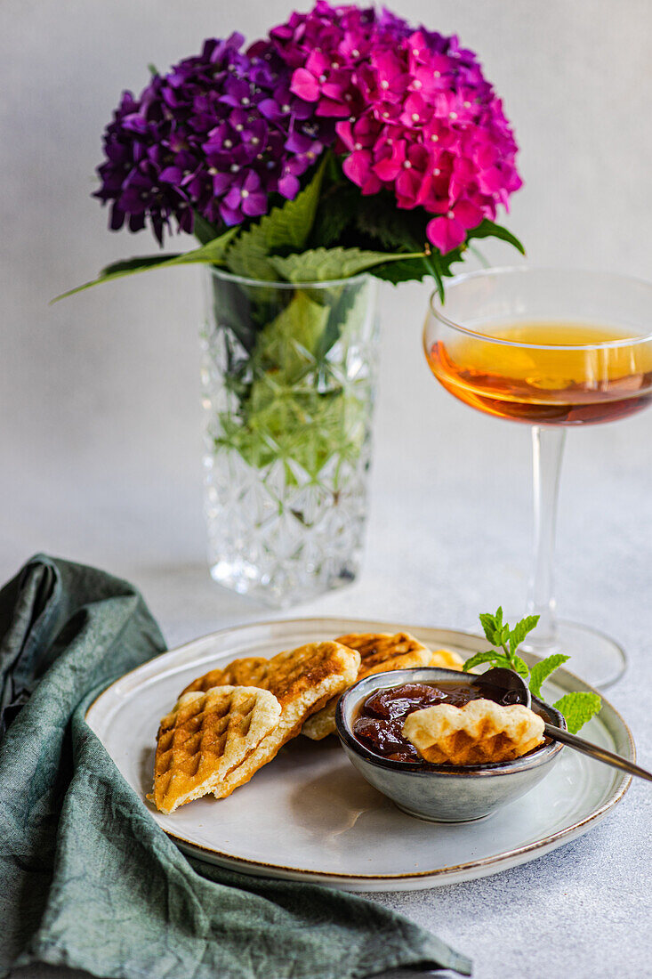 Appetitanregende süße Waffeln mit Pfirsichmarmelade auf rundem Tisch neben Glas mit Getränk im Garten mit blühenden lila Hortensienblüten