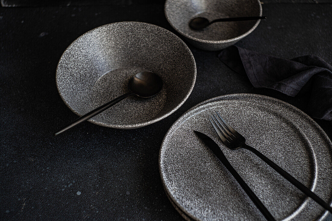 An elegant arrangement of dark stoneware dishes with sleek utensils, against a black backdrop, perfect for sophisticated dining settings.