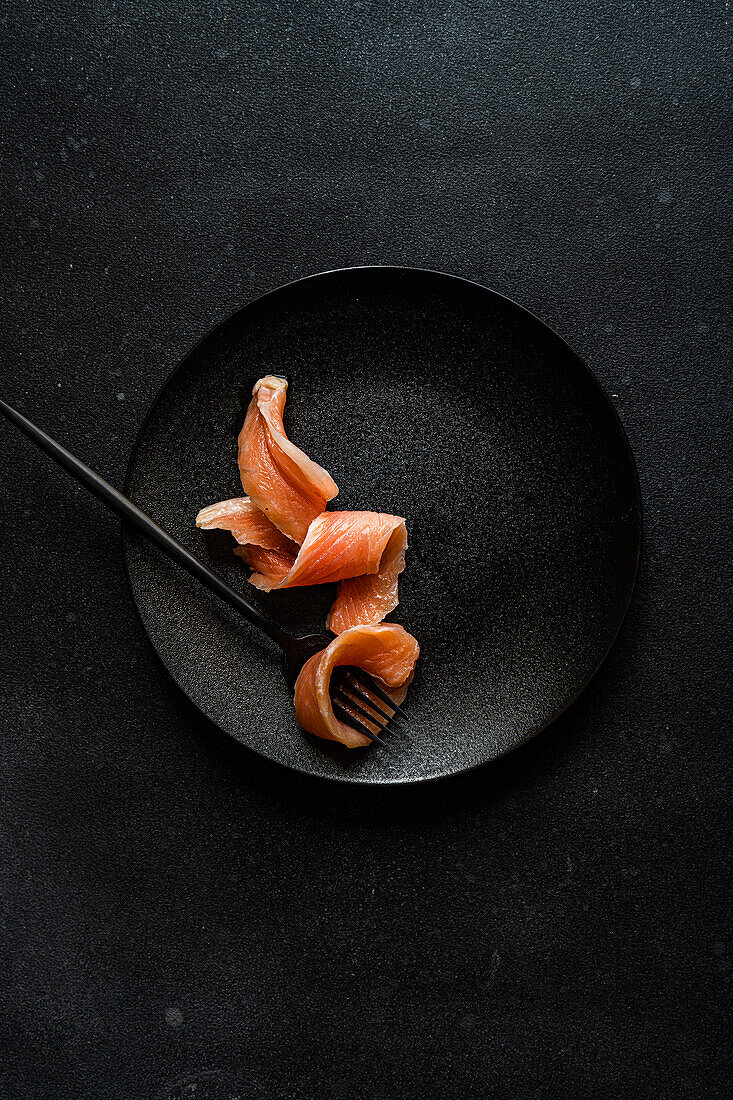 Draufsicht auf eine gesunde Lachsscheibe, serviert auf einem schwarzen Teller neben einer Gabel vor dunklem Hintergrund