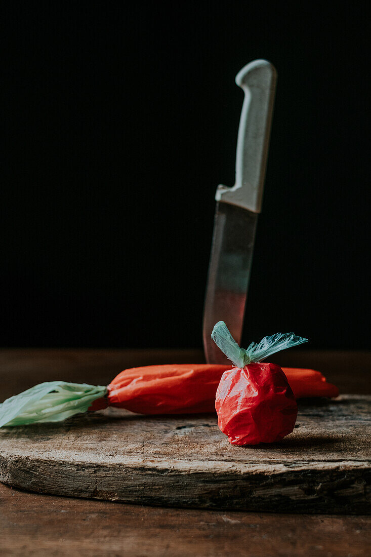 Plastic bags crafted to resemble a tomato and carrot alongside a kitchen knife on a rustic wooden chopping board