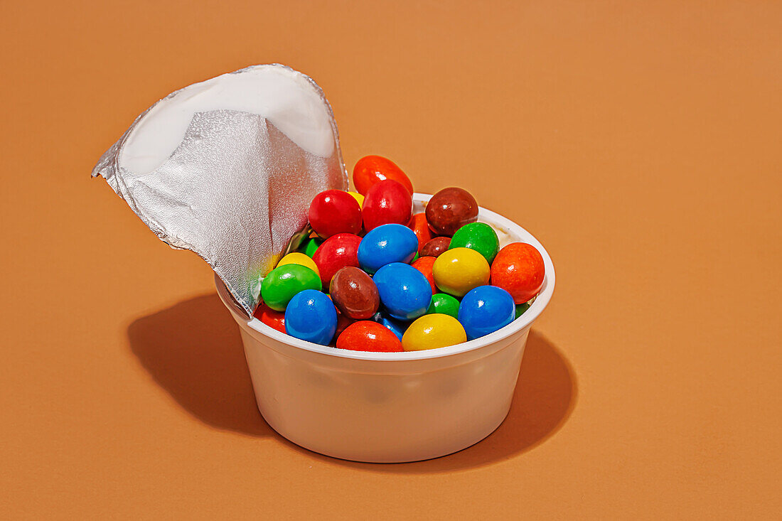 Nahaufnahme eines geöffneten Joghurtbehälters, gefüllt mit bunten Bonbons vor orangefarbenem Hintergrund