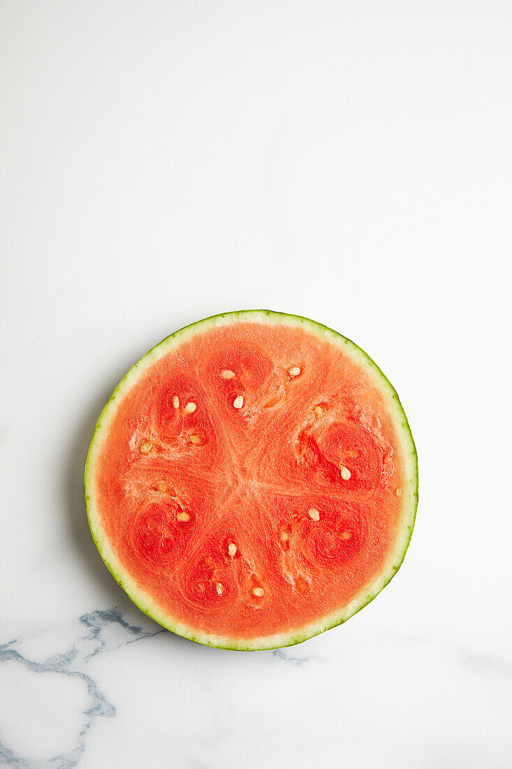 Draufsicht auf eine halb aufgeschnittene, reife, saftige Wassermelone mit Kernen, die auf einer weißen Keramikfläche im Tageslicht liegt