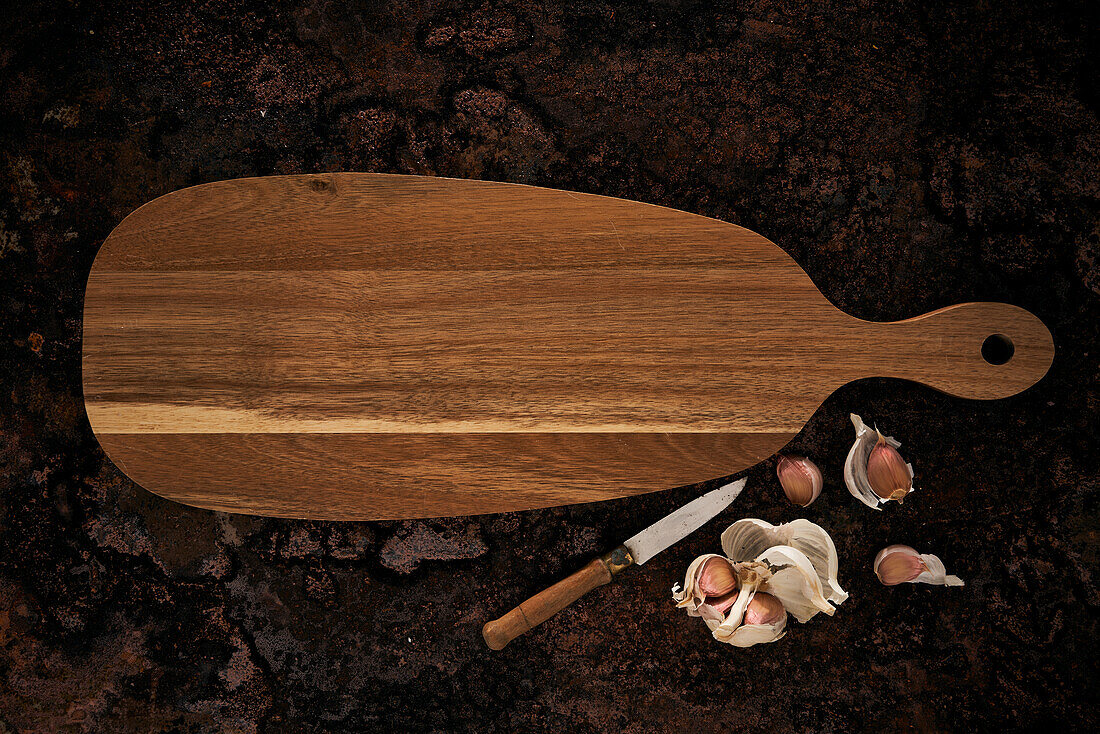 Top view of wooden oval shaped cutting board placed near knife and garlic on rough surface