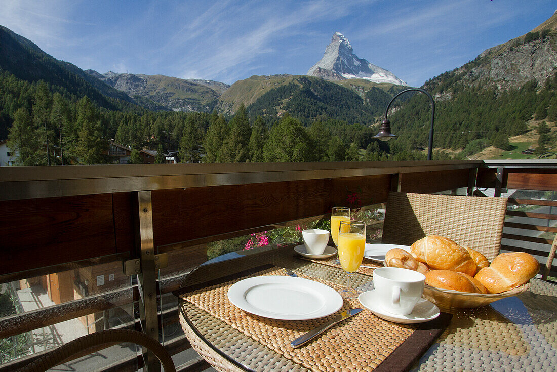 An outdoor dining setup overlooking the majestic Matterhorn mountain, with a spread of breakfast items including orange juice, croissants, and coffee surrounded by lush greenery