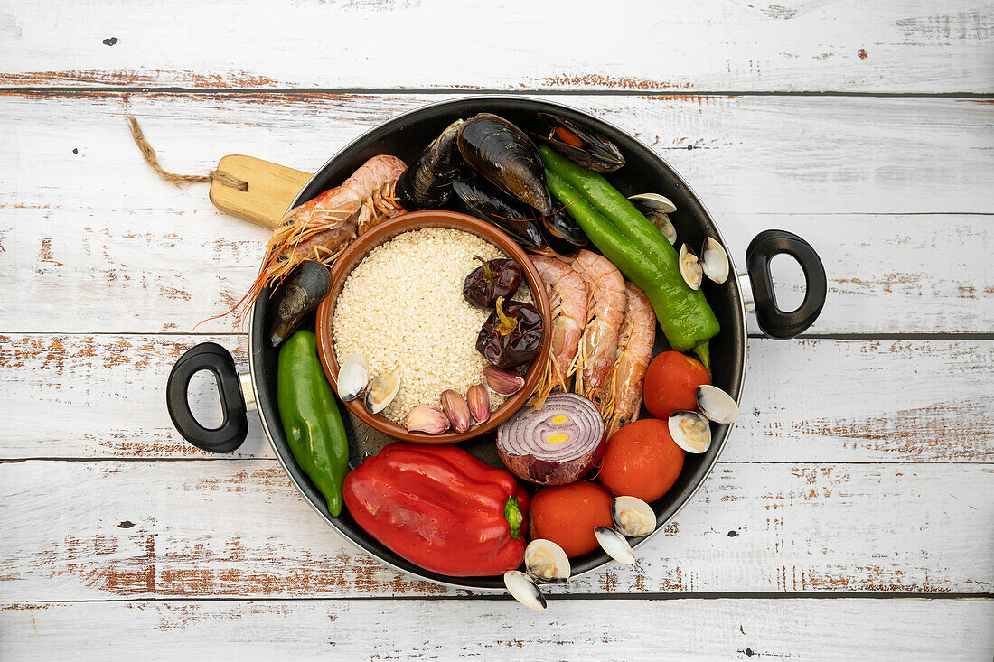 Blick von oben auf Reis in einer Schüssel mit verschiedenen rohen Meeresfrüchten, Gemüse und Gewürzen in einer Pfanne auf dem Tisch für die Zubereitung einer Paella in der Küche