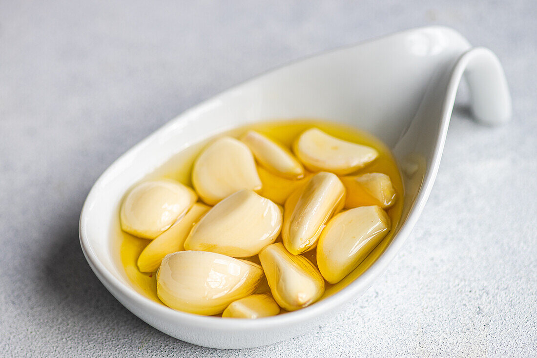 Closeup peeled and baked garlic cloves submerged in oil, showcased in a ceramic spoon against a light gray background