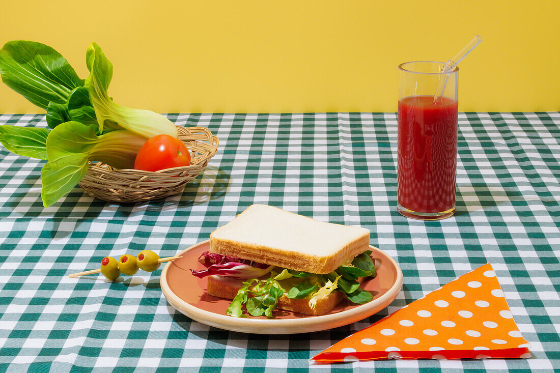 Appetitliches gesundes Sandwich mit frischem Salat auf einem Teller mit Oliven neben einem Glas Tomatensaft mit Glasstrohhalm auf einem karierten Tischtuch