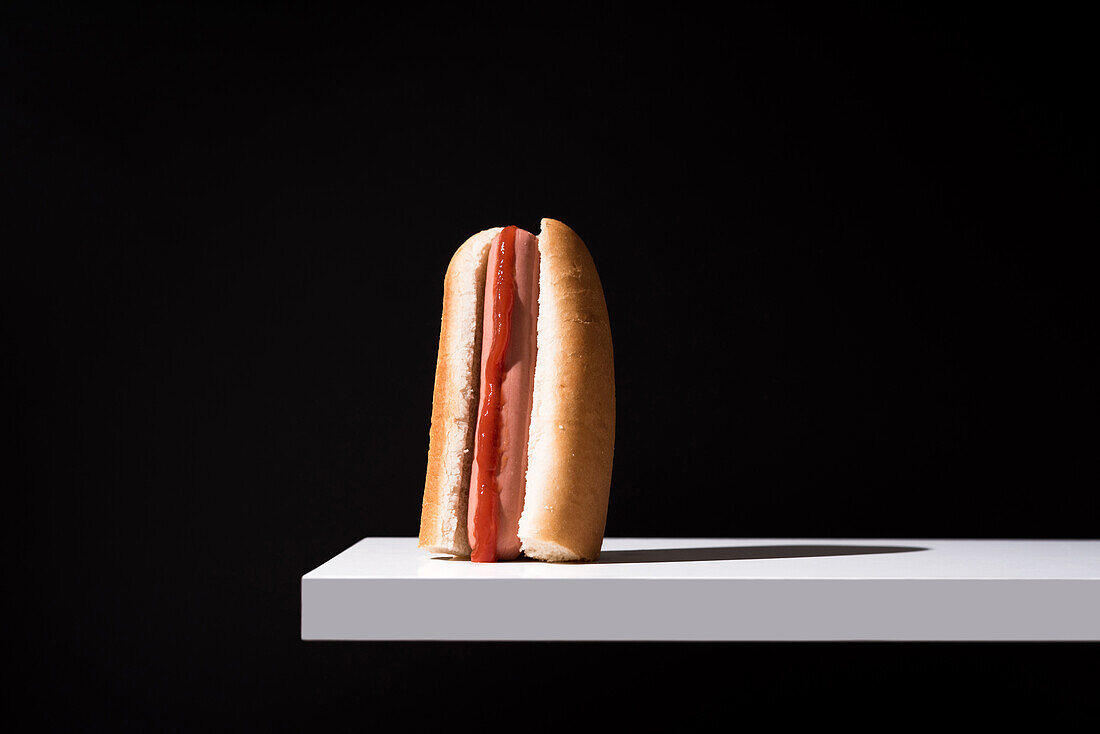 Appetitliches Brötchen mit Wurst und Ketchup auf einem weißen Holzbrett vor schwarzem Hintergrund im Studio