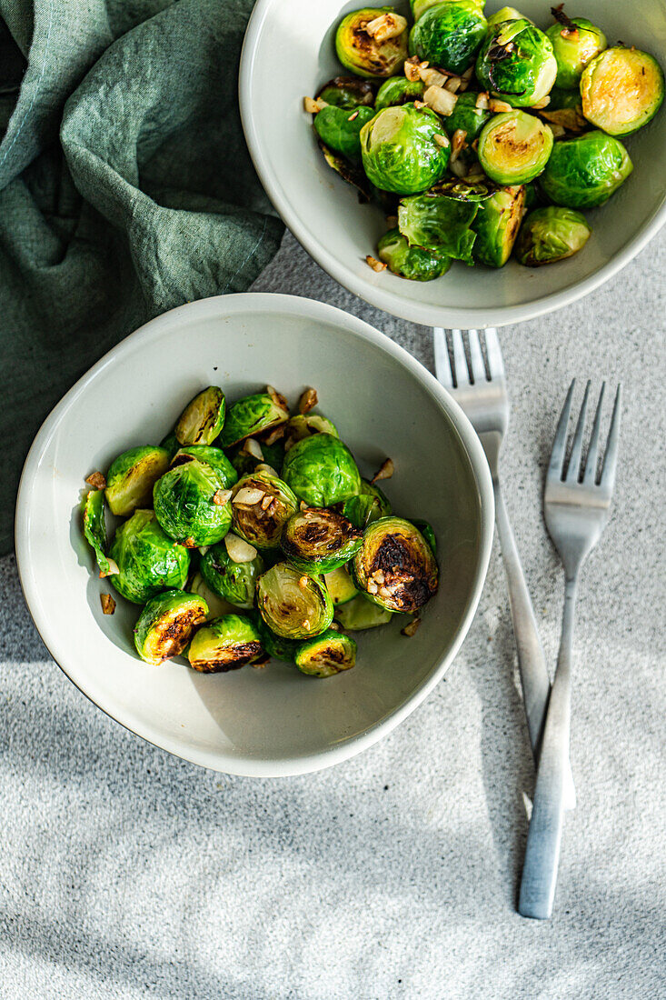 Top view of two bowls filled with barbecued Brussels sprouts flavored with garlic and spices, accompanied by silver forks on a textured tablecloth