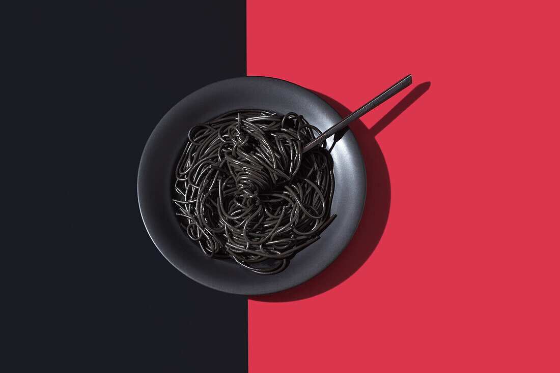 Blick von oben auf schwarze Spaghetti in einer Schüssel mit Gabel auf schwarzem und rotem Hintergrund