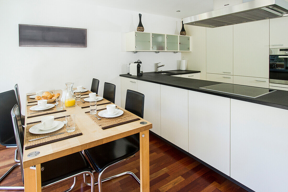 Moderne Küche mit weißen Schränken, gedecktem Esstisch und eleganten Geräten. Das warme Ambiente wird durch den Holzfußboden unterstrichen