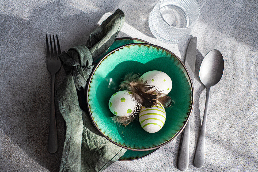 Top view of Easter table setting, showcasing a vibrant green ceramic plate with two decorative Easter eggs adorned with white and green patterns and delicate feathers, placed on gray surface between napkin and cutlery and glass of water