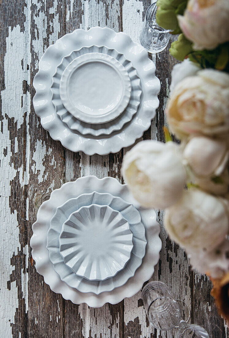 Draufsicht auf verschiedene weiße dekorative Keramikteller auf einem schäbigen Holztisch neben blühenden Blumen in einer Vase