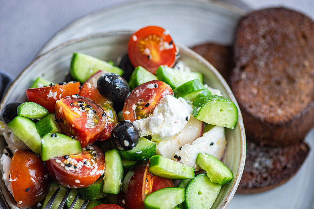 Closeup of ceramic bowl with tasty healthy salad, vegetables and bread placed with sliced cucumber and tomato with fabric over gray table