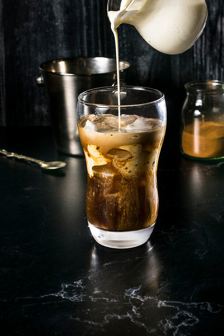 An enticing glass of Greek frappe mid-preparation, with a stream of milk being poured, creating a beautiful swirl in the combination of instant coffee and ice