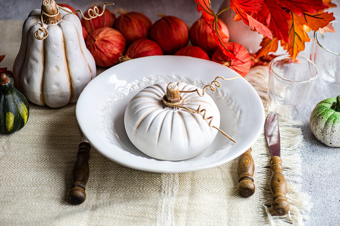 High angle of autumnal table setting with white pumpkin on plate placed on table near cutlery, glasses and red onions and leaves
