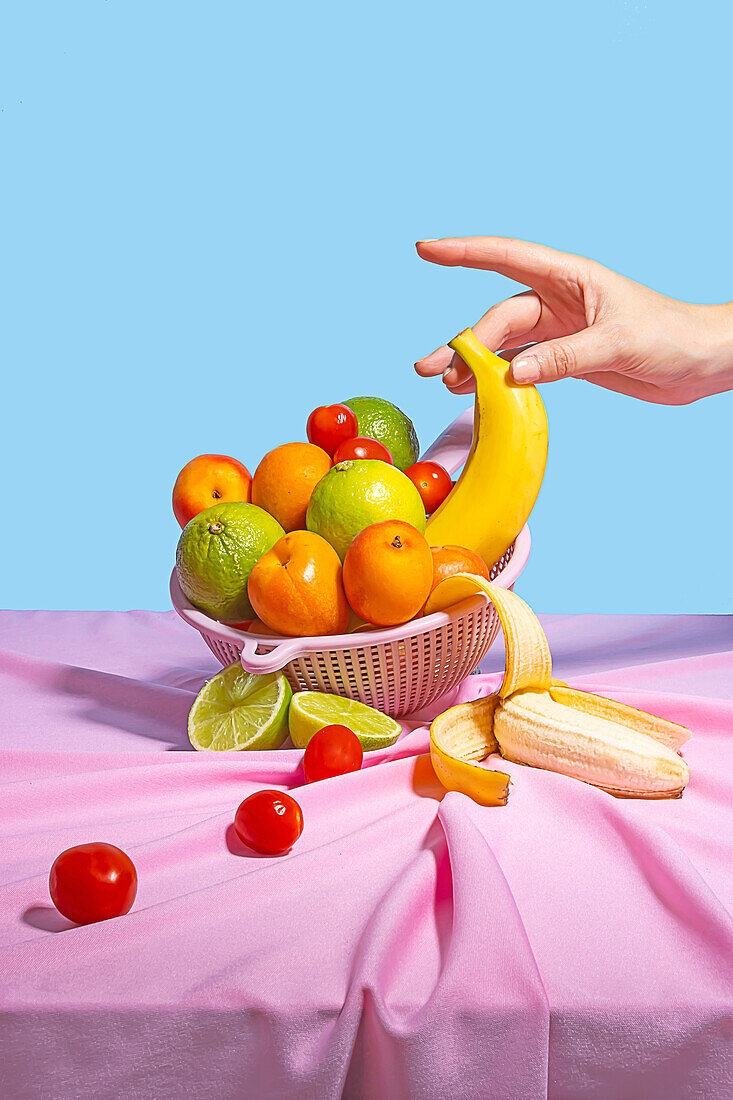 Anonyme Person, die eine Banane von einem Plastikständer mit frischem Obst auf einem Tisch mit einer rosa Tischdecke nimmt