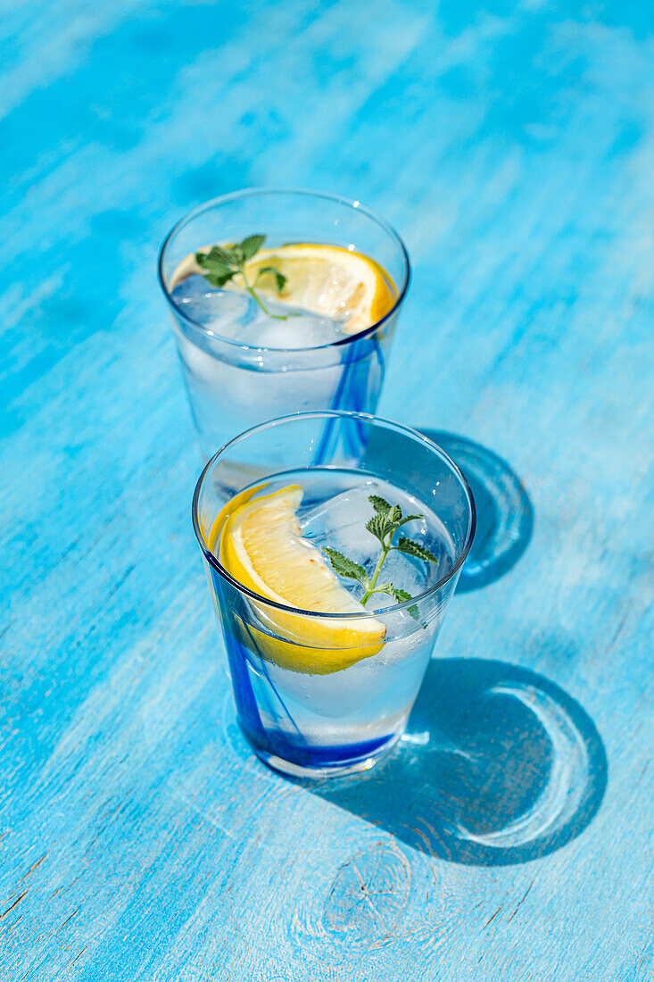 High angle of Summer cocktails with lemon vodka, slices of lemon and wild mint leaves served with ice placed on blue table