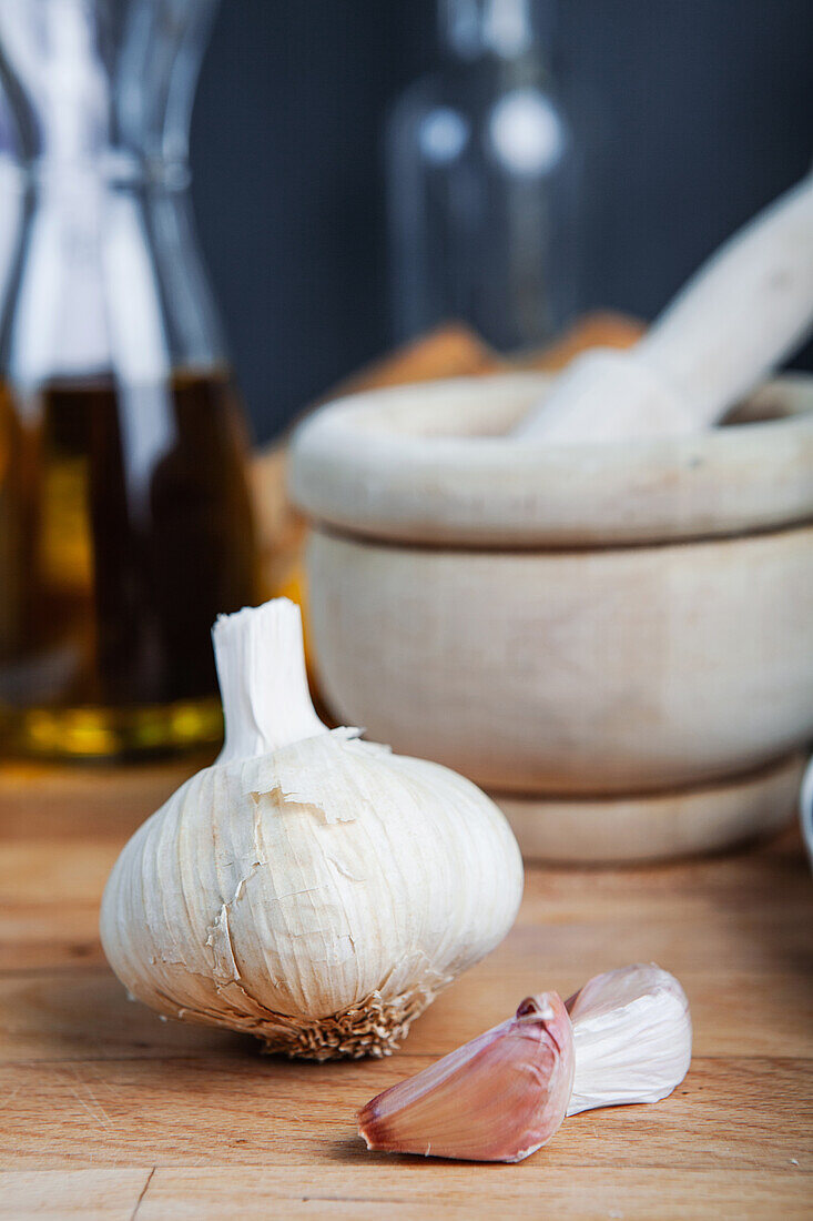 A close-up image featuring a whole garlic bulb and individual cloves with a mortar and kitchen utensils in the blurry background.