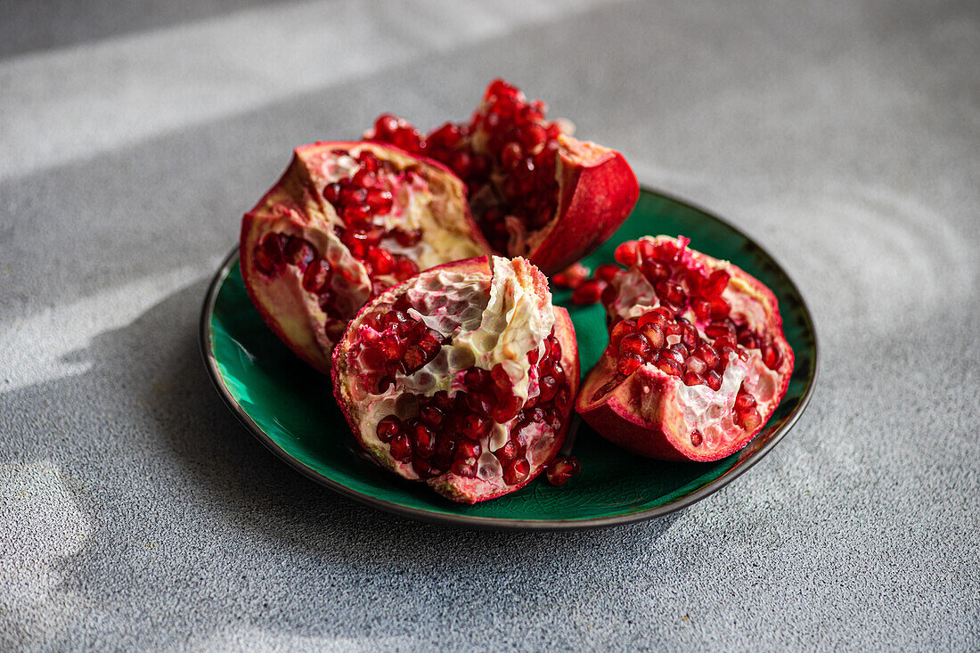 From above fresh pomegranate pieces with vibrant red seeds on a green ceramic plate set against a textured grey concrete background