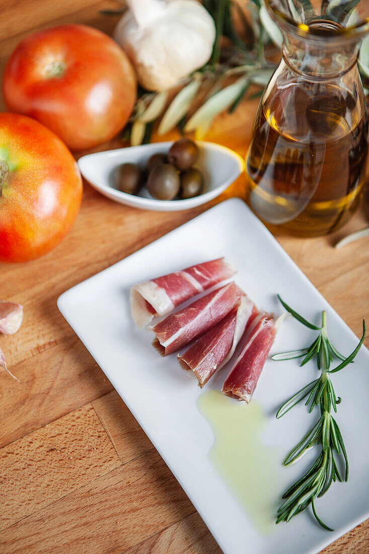 A wooden table showcases Mediterranean ingredients with a focus on sliced iberian ham, fresh tomatoes, garlic, olives, and a bottle of olive oil with rosemary.