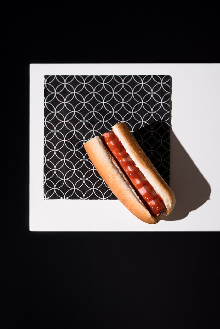 High angle of appetizing bun with sausage and ketchup served table mat over white wooden board against black background in studio